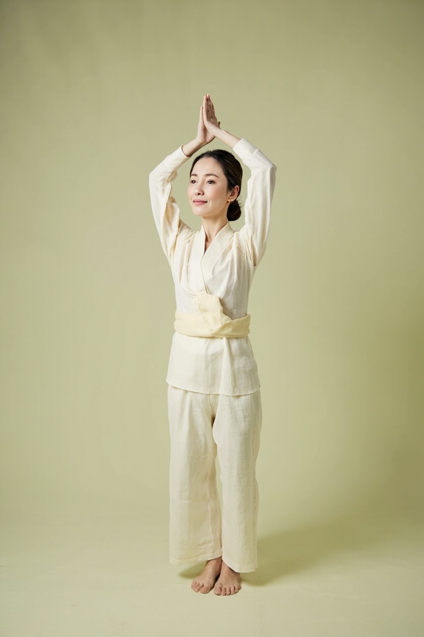 A calm woman in warm-cream linen practising a slow standing Ba Duan Jin qigong movement, soft natural light against a sage background