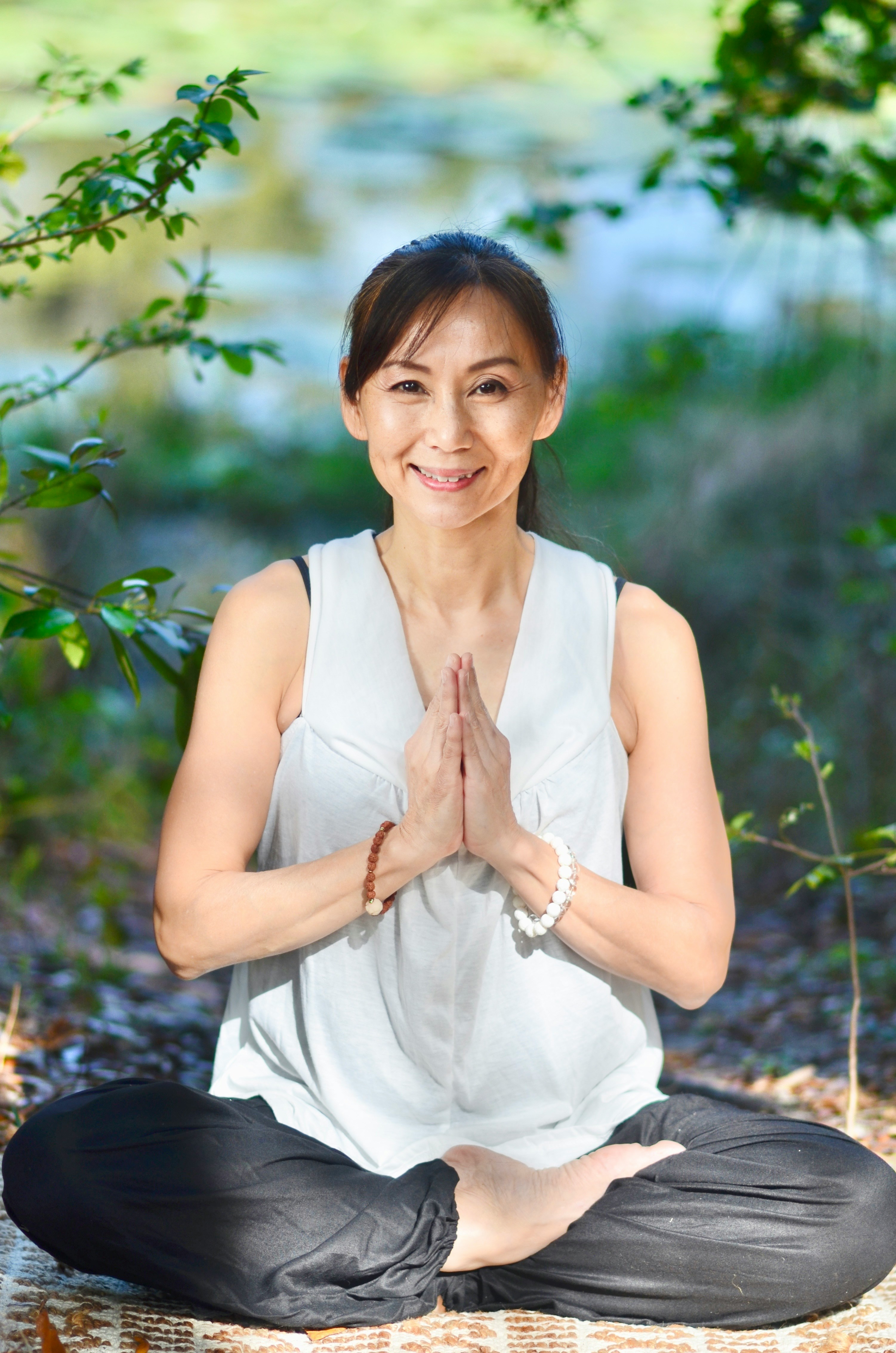 A woman seated in meditation outdoors among green trees, hands in prayer position — embodying the calm, grounded spirit of qigong practice
